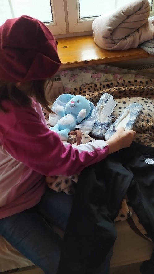 Na zdjęciu kobieta siedząca na łóżku, ubrana w spodnie koloru niebieskiego oraz bluzę w kolorze różowym, na głowie bordowa czapka. Obok na kocu leżą niebieskie kapcie i niebieski pluszowy króliczek. W tle okno z drewnianym parapetem